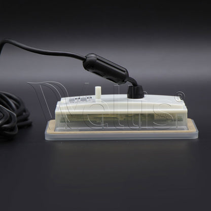 A small rectangular electronic device with a clear plastic cover, beige base, white top panel, and attached black power cord, photographed on a dark studio background.